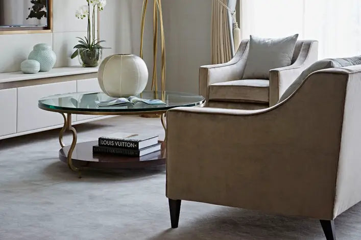 A modern living room scene featuring a beige upholstered armchair in the foreground, a matching armchair behind it, a round glass-top coffee table with gold legs, and a white sideboard with decorative vases and a potted plant.