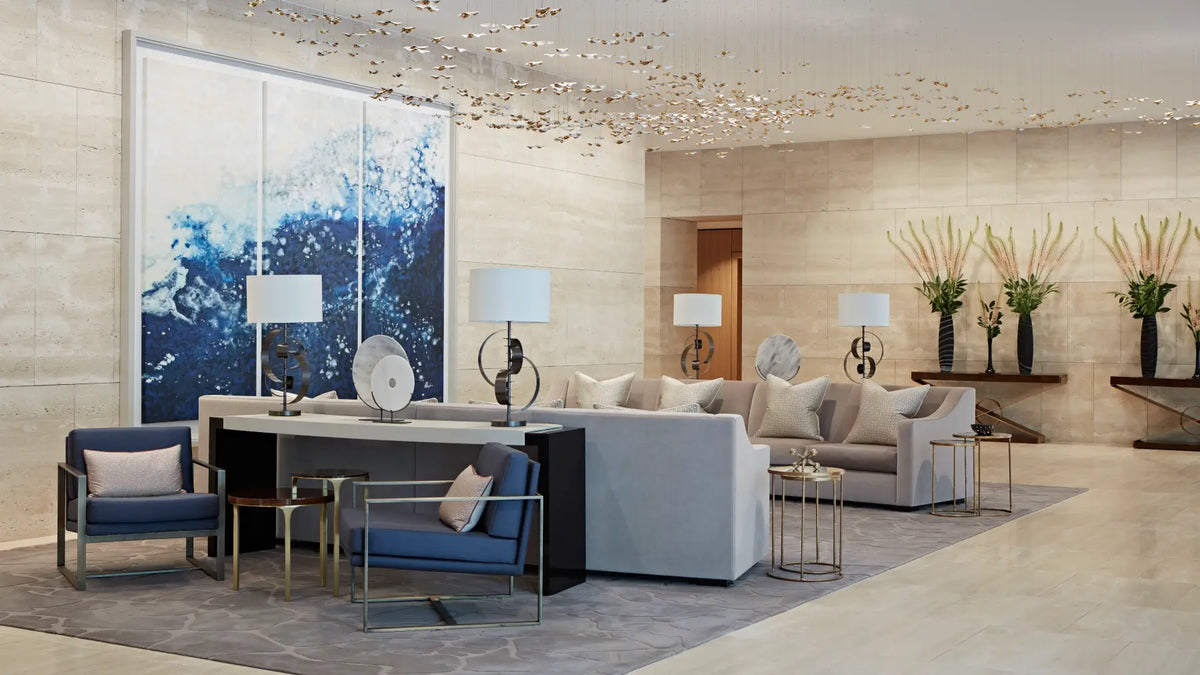 A modern, luxurious interior lounge space featuring a large abstract blue and white wall art piece, contemporary blue armchairs, a light gray sectional sofa, and a central dark wood console table with matching lamps.