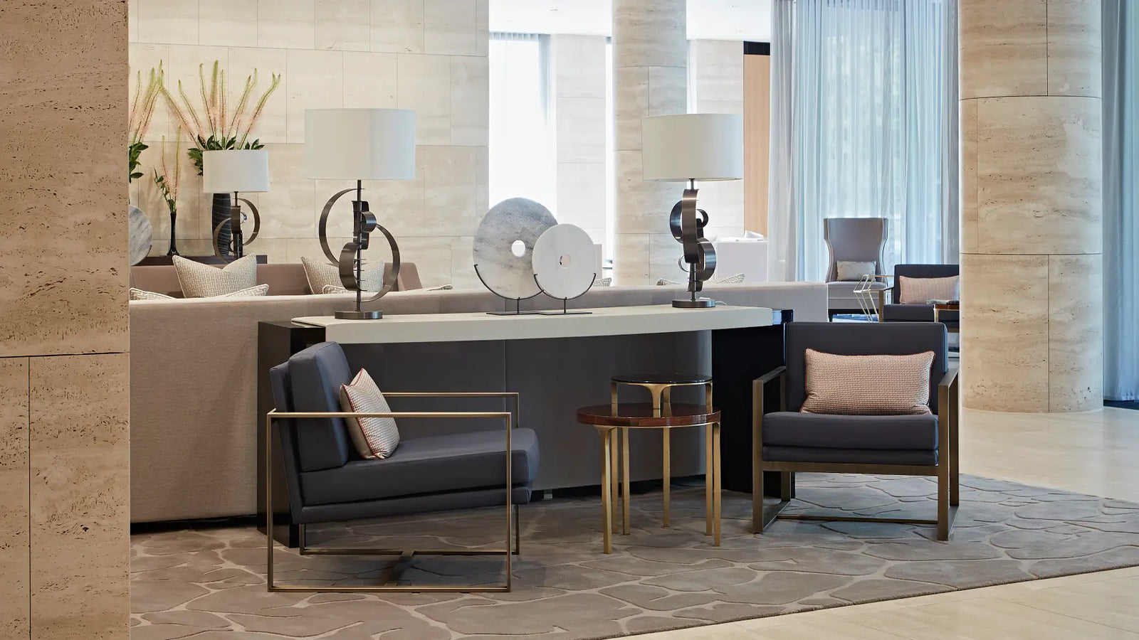 A modern lounge area featuring two dark gray armchairs with gold metal frames, light pink accent pillows, and a small round wooden side table between them, set on a patterned rug in a contemporary stone-walled interior.