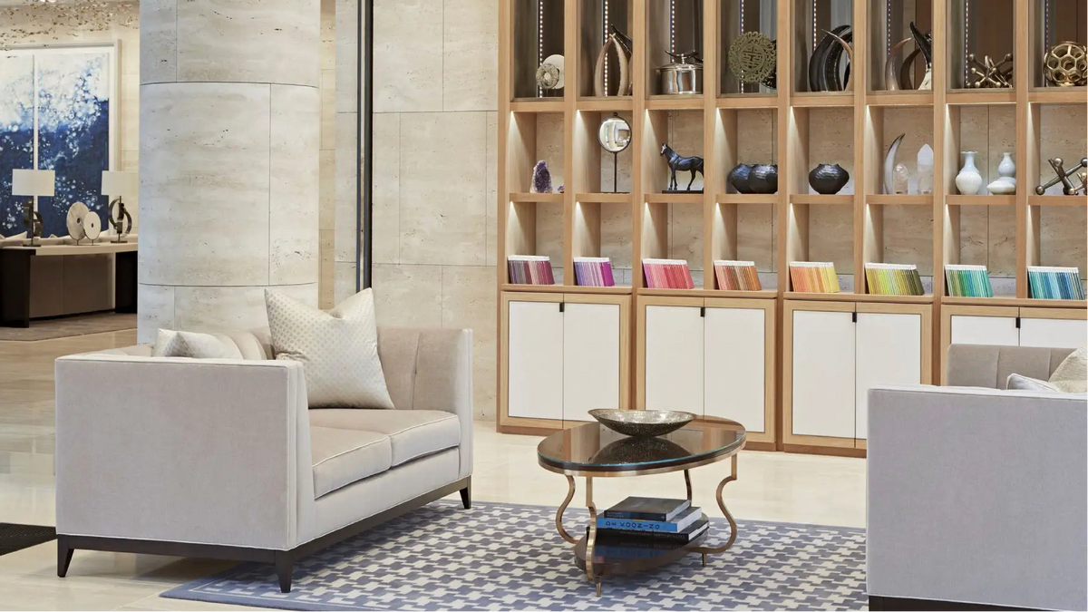 A modern living room setting featuring a light beige upholstered loveseat with white pillows, a round glass-top coffee table on a blue patterned rug, and a large wooden shelving unit in the background.