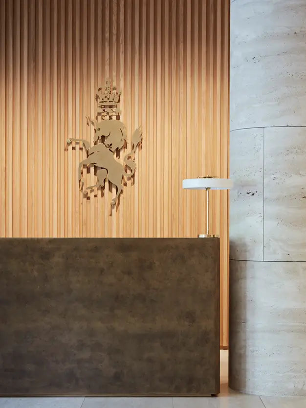 A modern interior scene with a dark textured reception desk in front of a warm wood slat wall featuring a metallic horse-and-crown emblem, next to a light stone column and a small table lamp.