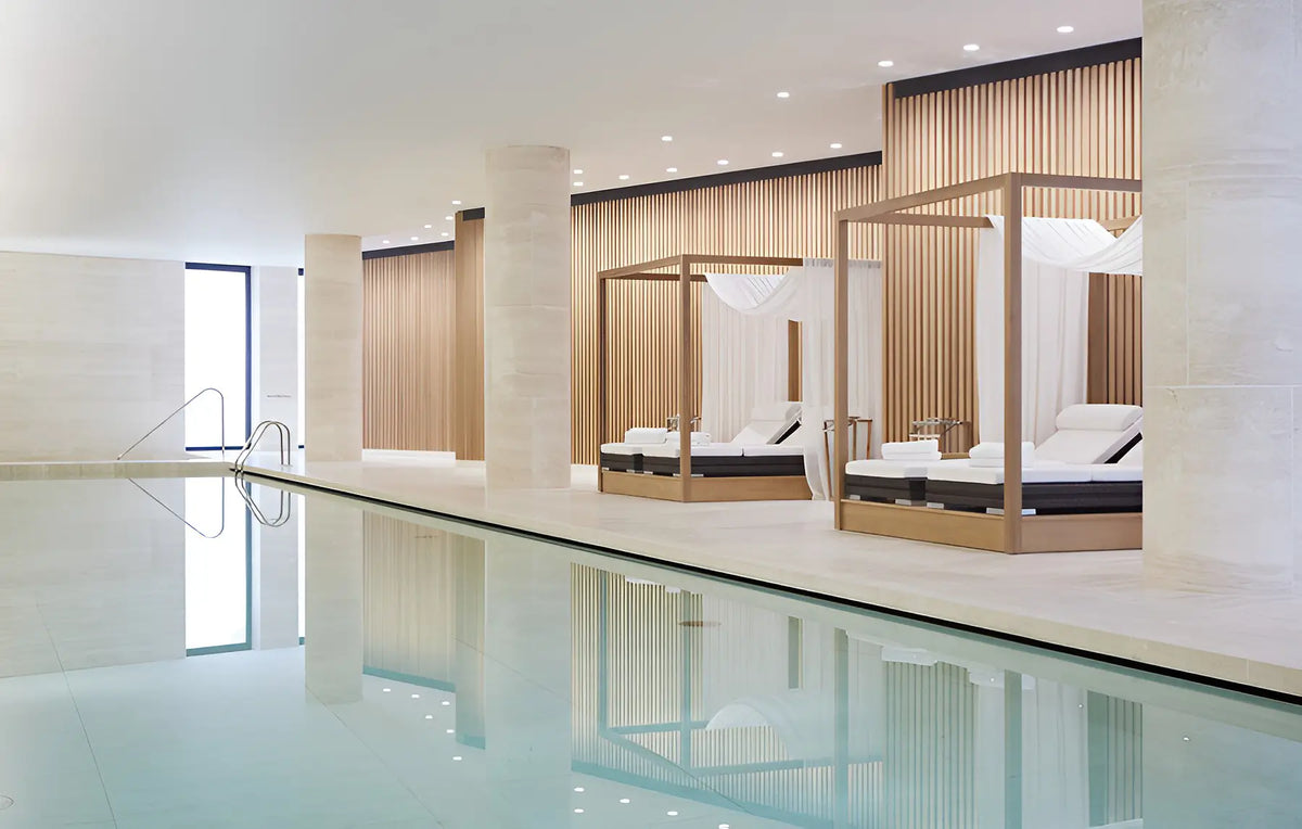 Luxury indoor poolside lounge area with two wooden canopy daybeds on a light stone terrace, reflecting in the calm water.