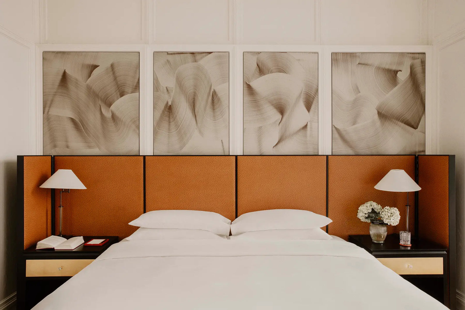 A modern bedroom setup featuring a bed with white linens, dark wood nightstands with white table lamps, and a prominent orange-brown segmented headboard. Above the headboard are four matching abstract canvas prints in beige and brown tones.