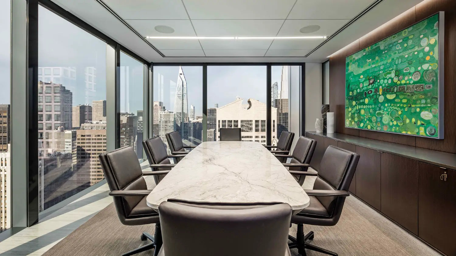 A modern conference room with a long marble-top meeting table, multiple dark gray leather chairs, large floor-to-ceiling windows overlooking a city skyline, and a large green abstract wall art piece on the right wall.