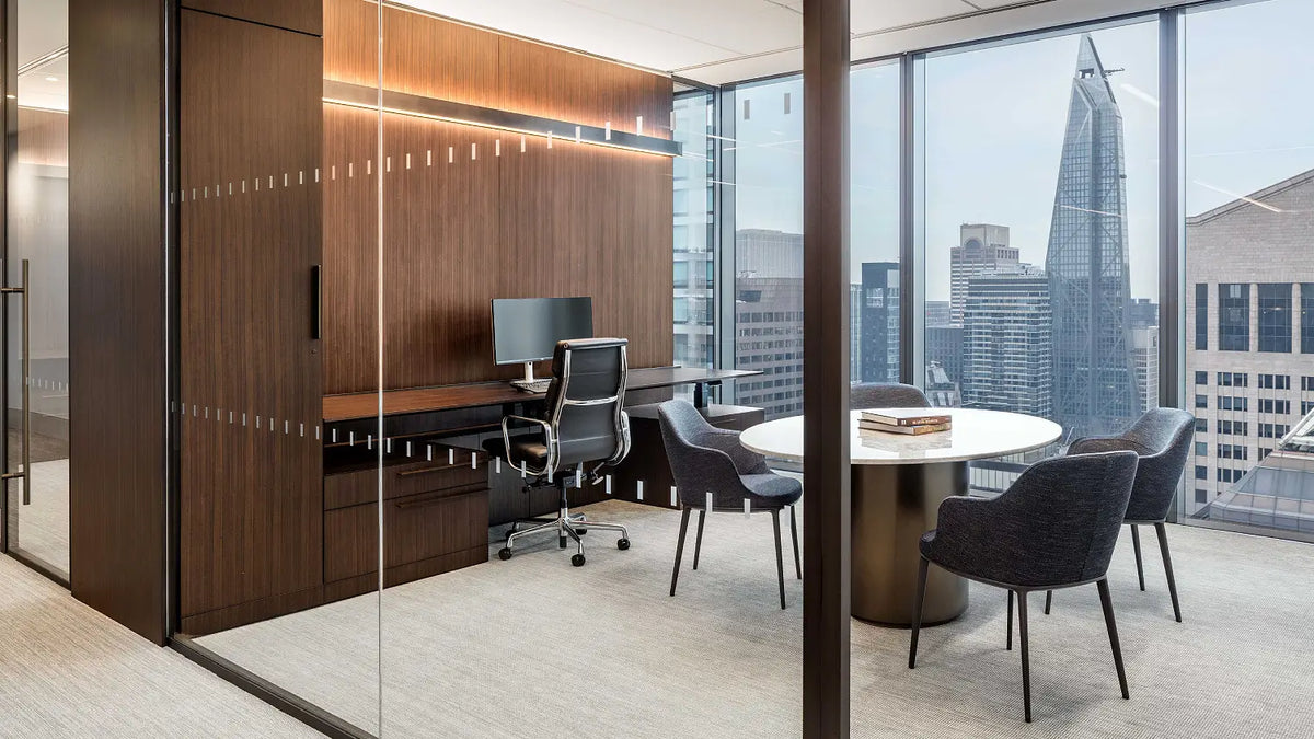 A modern glass-walled office meeting room with a round white table, dark gray chairs, a desk setup, and large windows overlooking a city skyline.