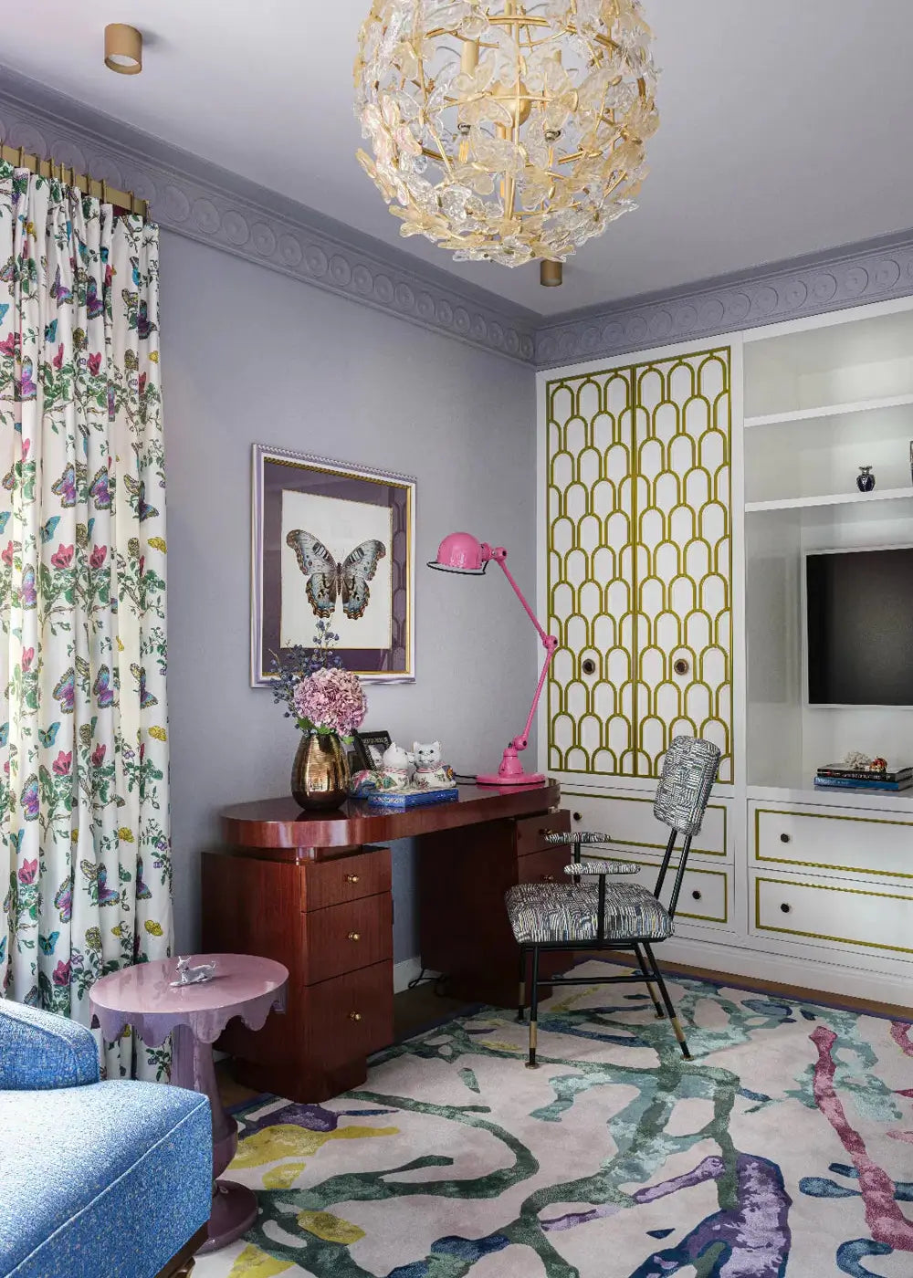 A styled home office or study room with a large crystal chandelier, a pink desk lamp, a floral curtain, a patterned rug, and built-in shelving with gold-accented paneling.