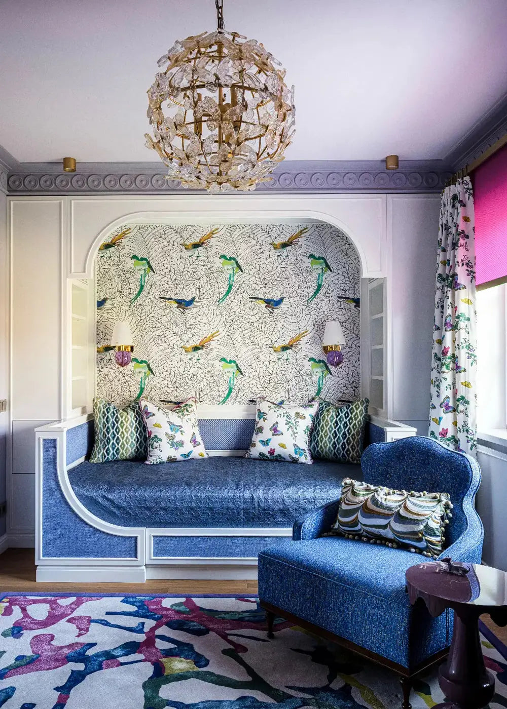 A styled interior room featuring a blue upholstered daybed with patterned pillows, a matching blue armchair, a large abstract area rug, a decorative wallpapered arched wall panel, and a hanging shell chandelier.