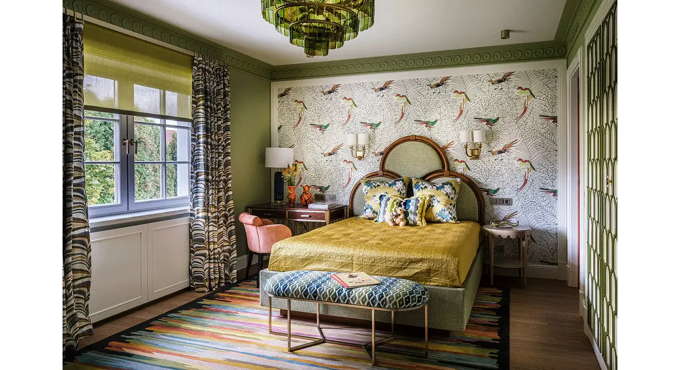 A styled bedroom interior featuring a bed with a yellow quilt, patterned pillows, a patterned bench, a striped rug, olive green walls, and a decorative wallpapered accent wall.