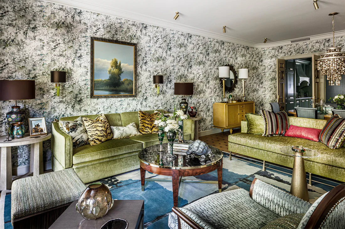 A luxurious living room featuring olive green velvet sofas, a matching armchair, a round wooden coffee table, and a blue patterned rug, all set against a black and white floral wallpapered wall.