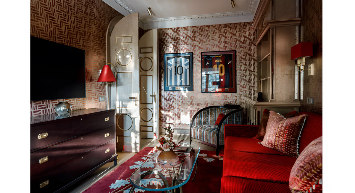 A luxury living room interior featuring a vibrant red sofa with patterned cushions, a matching red area rug, a dark wood chest, and textured geometric wallpaper.