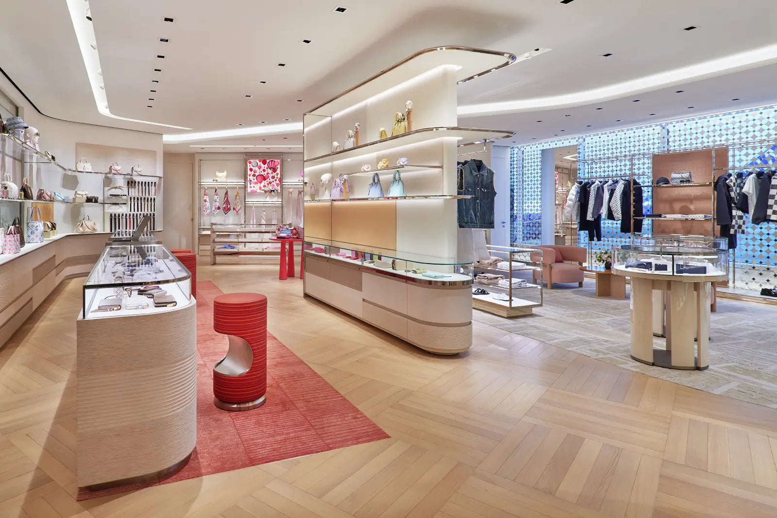 A luxury boutique interior with light wood flooring, curved beige display counters, glass cases, clothing racks, and a red rug. No people are visible.