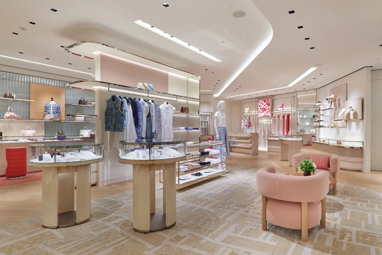 A luxury boutique interior with clothing racks, display tables, pink upholstered seating, and warm neutral flooring under bright recessed lighting.