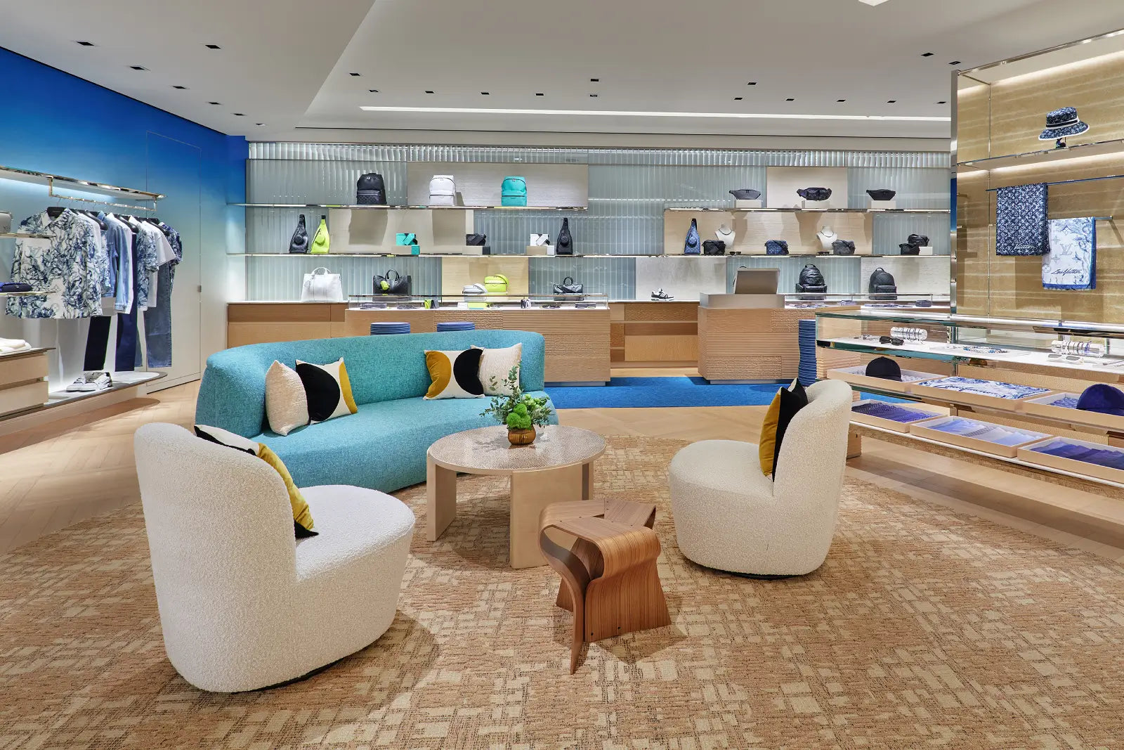 A luxury retail interior with a turquoise sofa, two cream boucle armchairs, a round wooden coffee table, and a small wooden stool on a patterned beige carpet. Shelves and clothing racks display merchandise in the background.