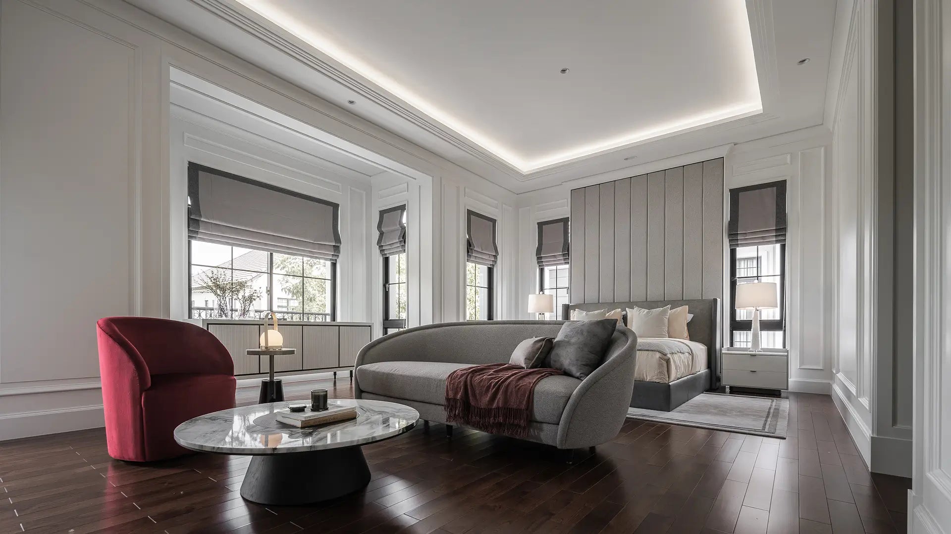 A luxury modern living room featuring a curved gray sofa, a deep red armchair, a round glass-top coffee table, and a bed in the background, set in a bright white room with dark wood floors.