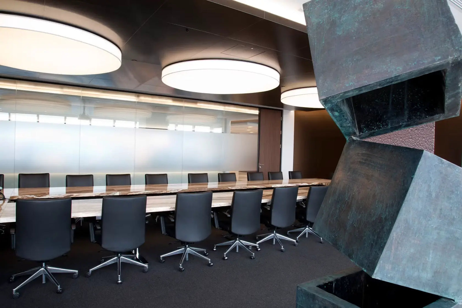 A modern conference room with a long rectangular table, multiple black swivel chairs, large circular ceiling lights, frosted glass windows, and a large abstract metal sculpture in the foreground.