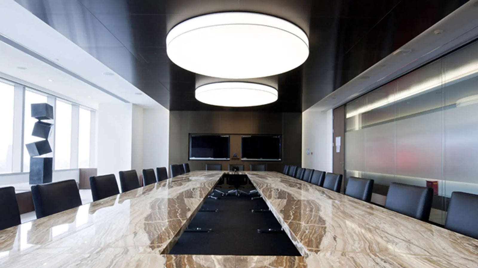 A modern conference room with a long polished marble boardroom table, black chairs, two large circular ceiling lights, and wall-mounted screens.