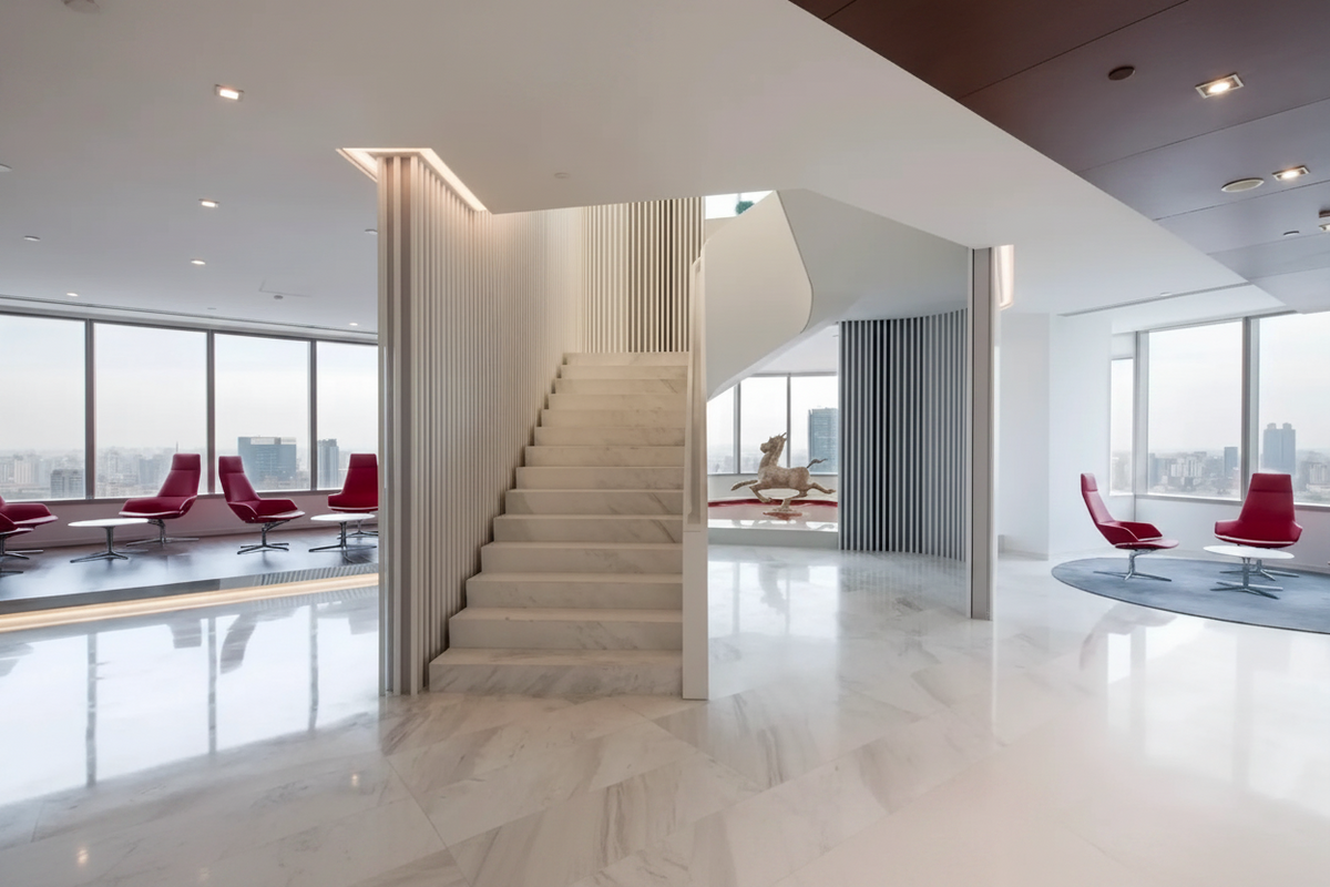 A modern interior lobby with a central marble staircase, large floor-to-ceiling windows, and red lounge chairs in a bright, contemporary office space.