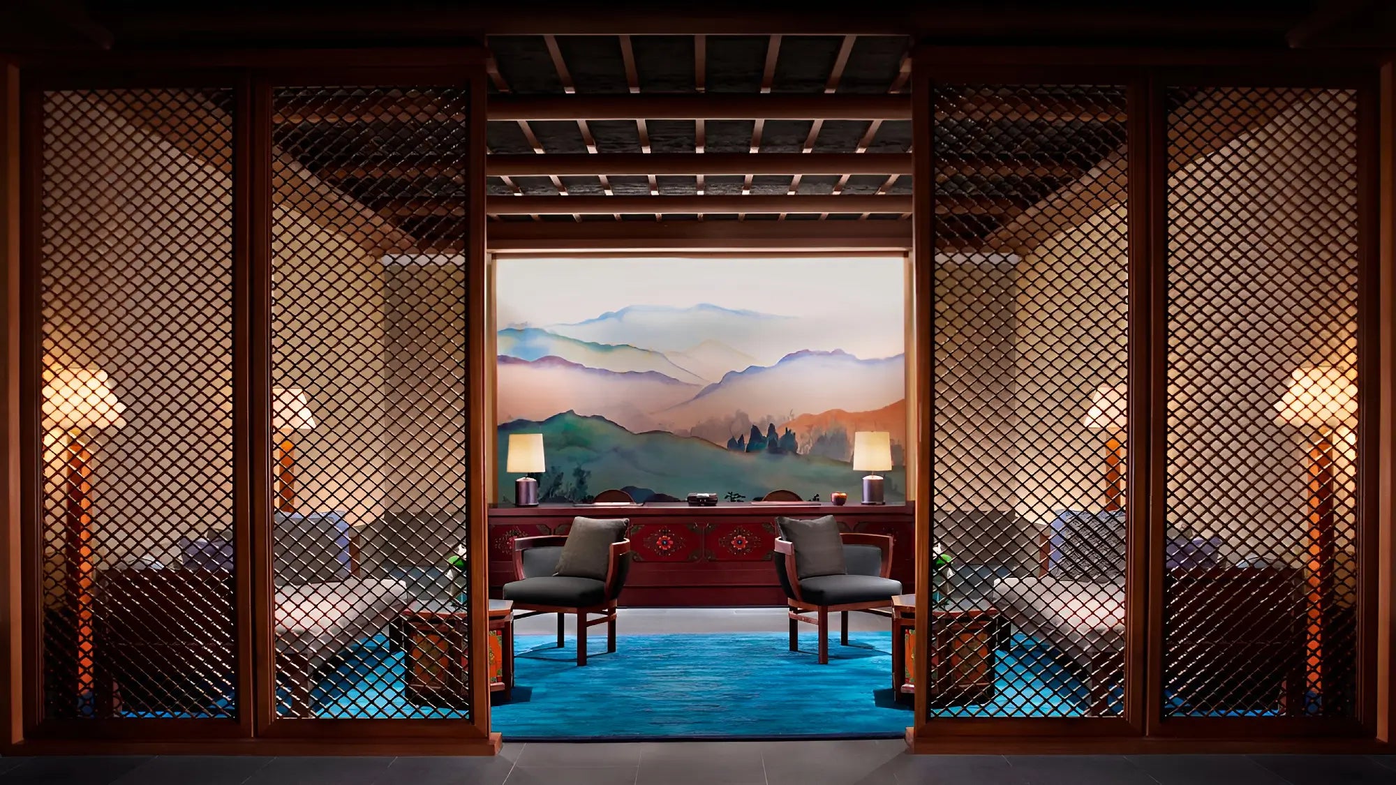A luxury interior lounge area featuring a central red wooden desk, two gray armchairs, and two upholstered sofas behind decorative lattice screens. The space is illuminated by warm floor lamps, set on a bright blue rug in front of a large scenic wall mural of mountains.