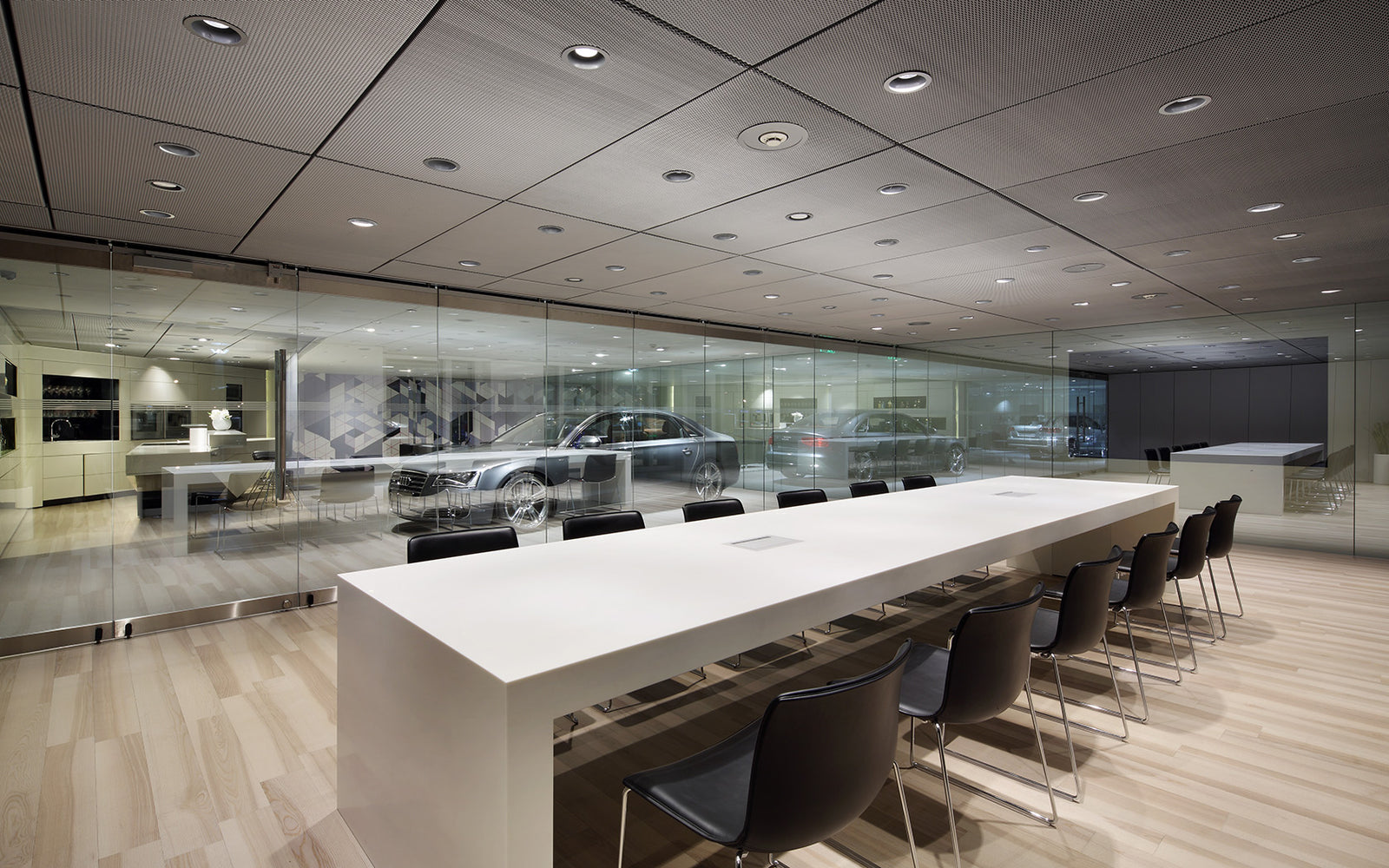 Modern showroom with a long white meeting table and chairs, glass walls, and cars displayed in the background.