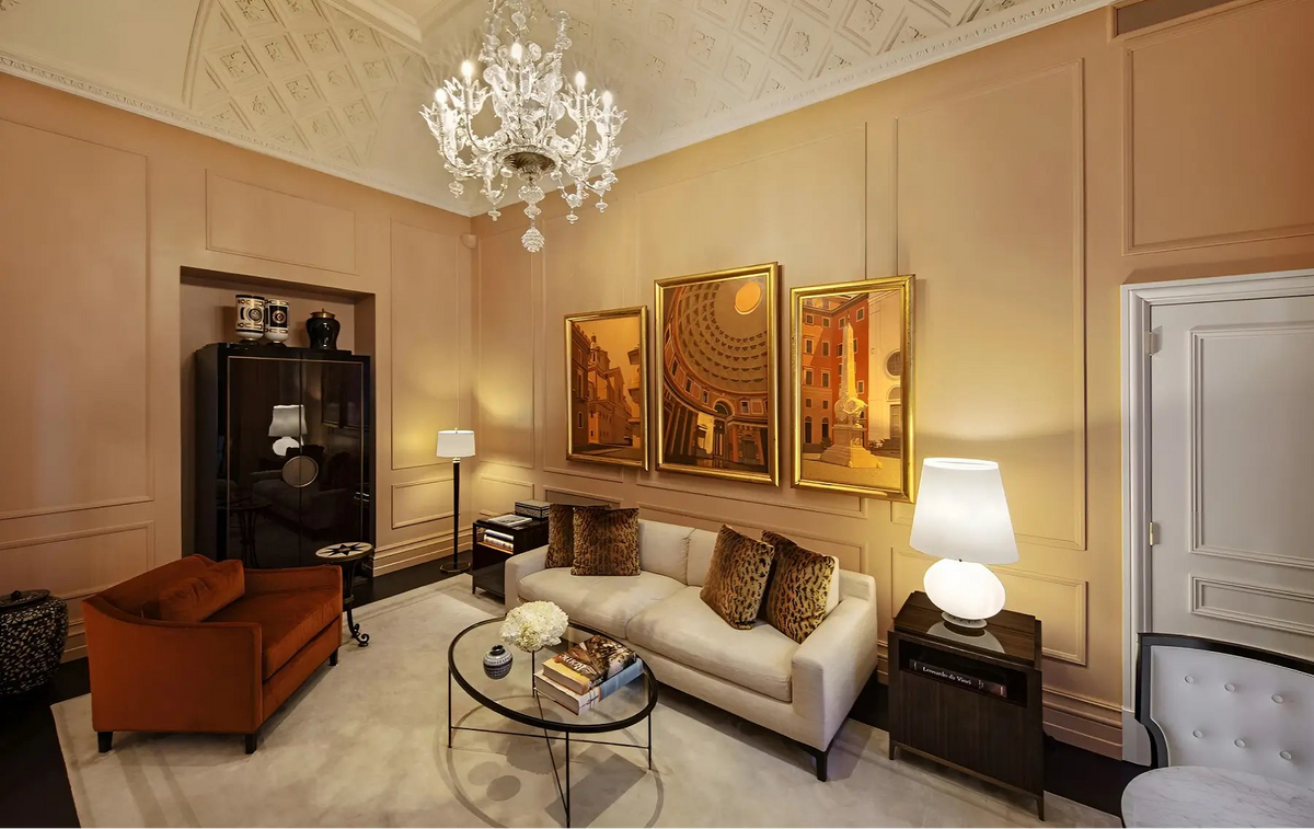 A luxury living room featuring a cream sofa with leopard-print pillows, a rust-orange armchair, a glass coffee table, and three gold-framed paintings on a beige paneled wall under a decorative coffered ceiling and crystal chandelier.