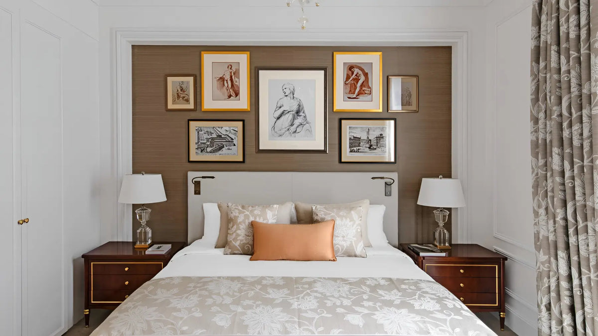A styled bedroom interior featuring a bed with white linens and a peach accent pillow, set against a brown accent wall decorated with multiple framed art prints. The room includes matching wooden nightstands, table lamps, and patterned curtains.