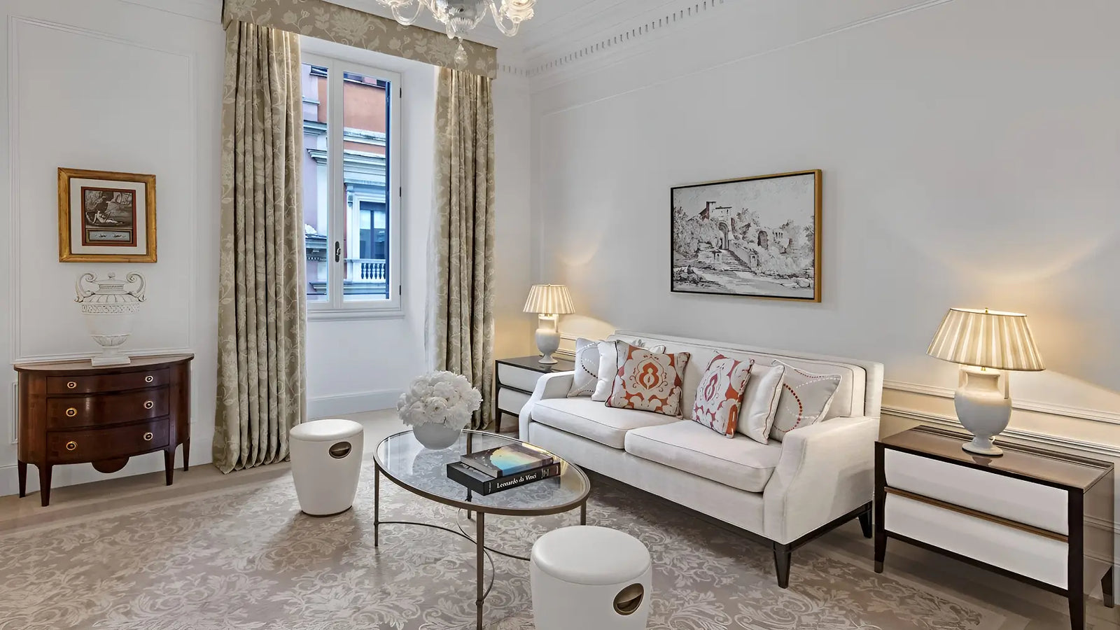 A styled living room interior featuring a white upholstered sofa with patterned pillows, a round glass-top coffee table, two white cylindrical stools, matching side tables with lit lamps, a dark wood cabinet, and beige curtains by a window.