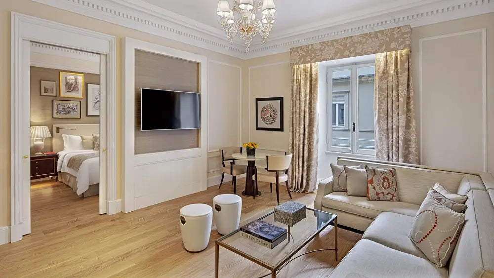 A luxury modern living room featuring a cream-colored sectional sofa, a glass-top coffee table, two white cylindrical stools, a built-in television on a wall, and an adjacent bedroom visible through an open doorway. The room has light wood floors, beige walls with white trim, and patterned beige curtains.