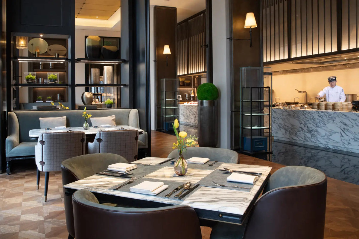 A luxury restaurant interior with a marble-topped dining table set with chairs, upholstered seating, shelving, and a chef visible in the background kitchen area.