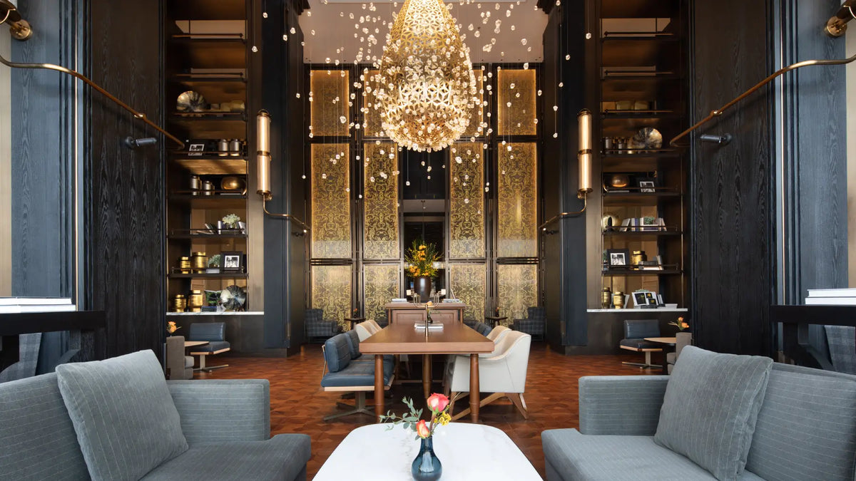 A luxury modern lounge or restaurant interior with dark wood shelving, a large glowing chandelier, a central dining table, gray upholstered seating, and warm ambient lighting.