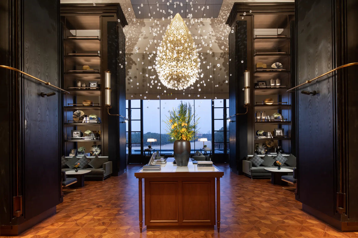 A grand interior space with dark wood shelving on both sides, a central wooden table with a floral arrangement, and a large glowing crystal chandelier hanging from a mirrored ceiling. Floor-to-ceiling windows in the back show an outdoor view at dusk.