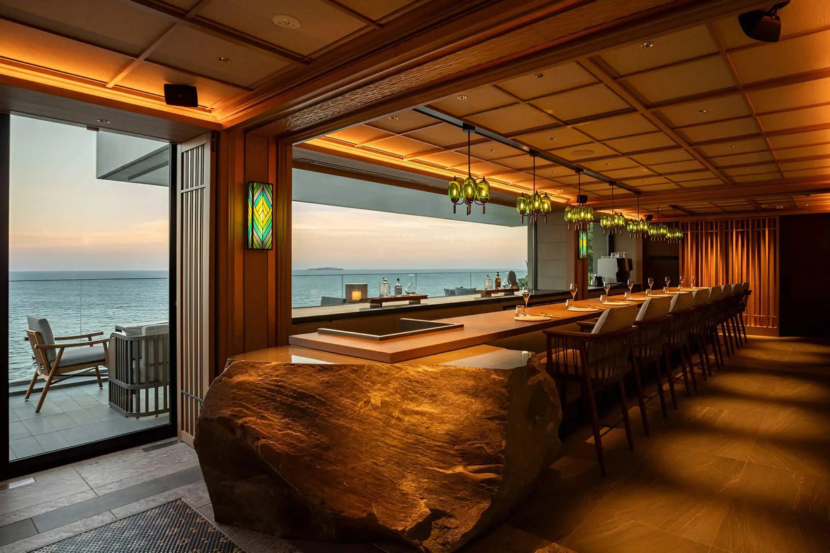 A luxurious modern bar and lounge interior with a long wooden bar, a large natural stone bar front, warm wood paneling, hanging glass pendant lights, and large windows overlooking the ocean at sunset.