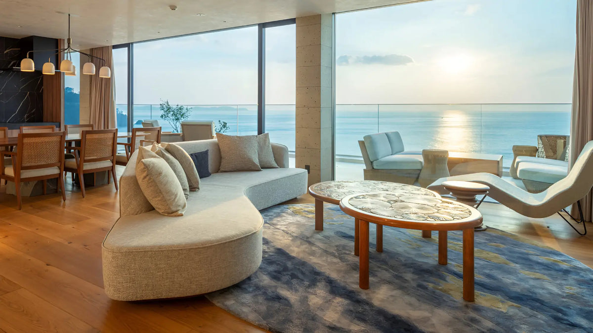 A modern living room with a curved beige sofa, round wooden coffee tables, and a large blue patterned rug, all set against floor-to-ceiling windows overlooking a calm ocean at sunset.