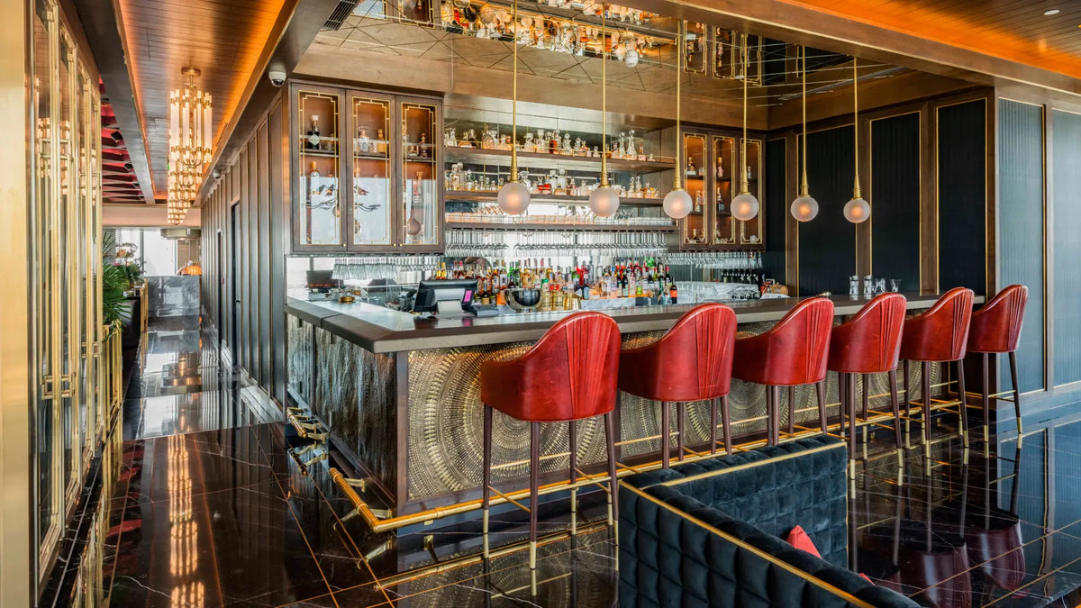 A luxury bar interior with a long counter, red velvet bar stools with gold legs, mirrored wood shelving filled with bottles, and warm ambient lighting over a dark polished floor.
