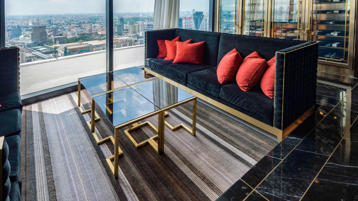 A luxury modern living room setup featuring a black velvet sofa with gold trim and red accent pillows, paired with matching geometric gold-framed glass coffee tables on a striped gray rug. The room is situated in a high-rise building with a panoramic city view through floor-to-ceiling windows.