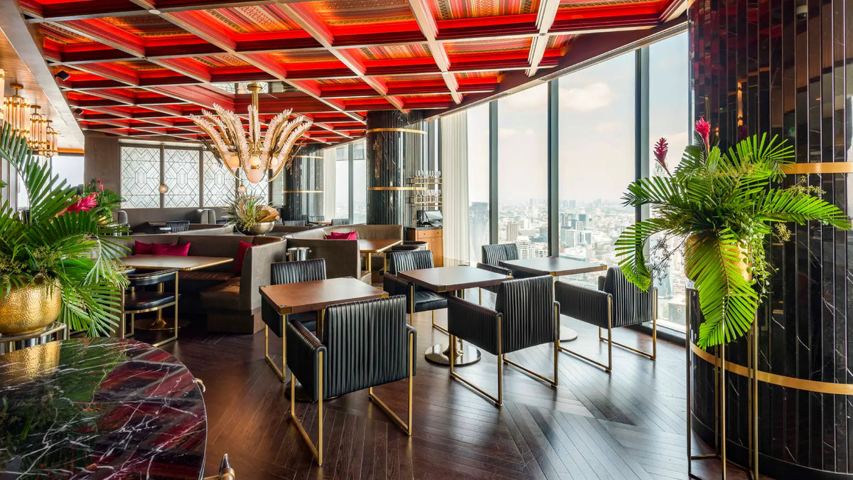 A luxury restaurant interior with a high ceiling featuring red glass panels, dark wood floors, black ribbed chairs with gold legs, and large windows overlooking a city skyline. Decorative potted plants and modern bar seating are visible throughout the space.