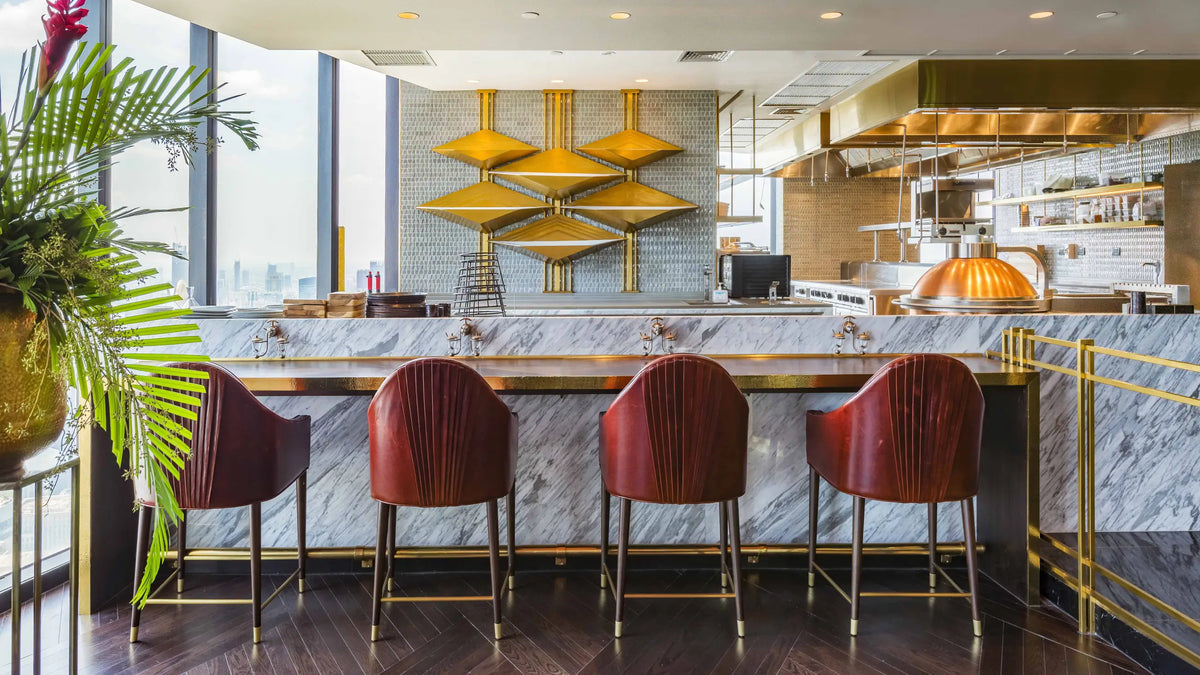 A modern interior scene with a marble-topped bar counter, four red upholstered bar stools with gold legs, a visible kitchen in the background, and large windows with a city view.