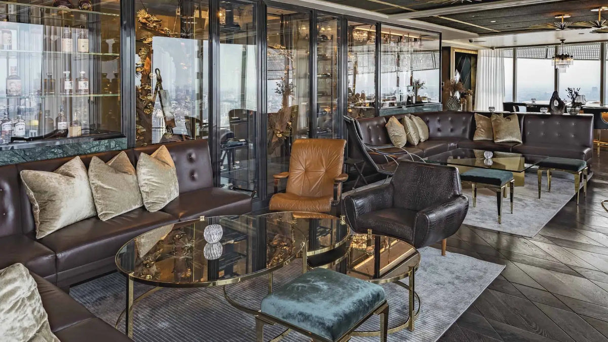 A luxury modern living room with dark brown leather sofas, a tan leather accent chair, a round glass-top coffee table with gold legs, and a teal velvet ottoman, set against a background of glass display cabinets and a city skyline.