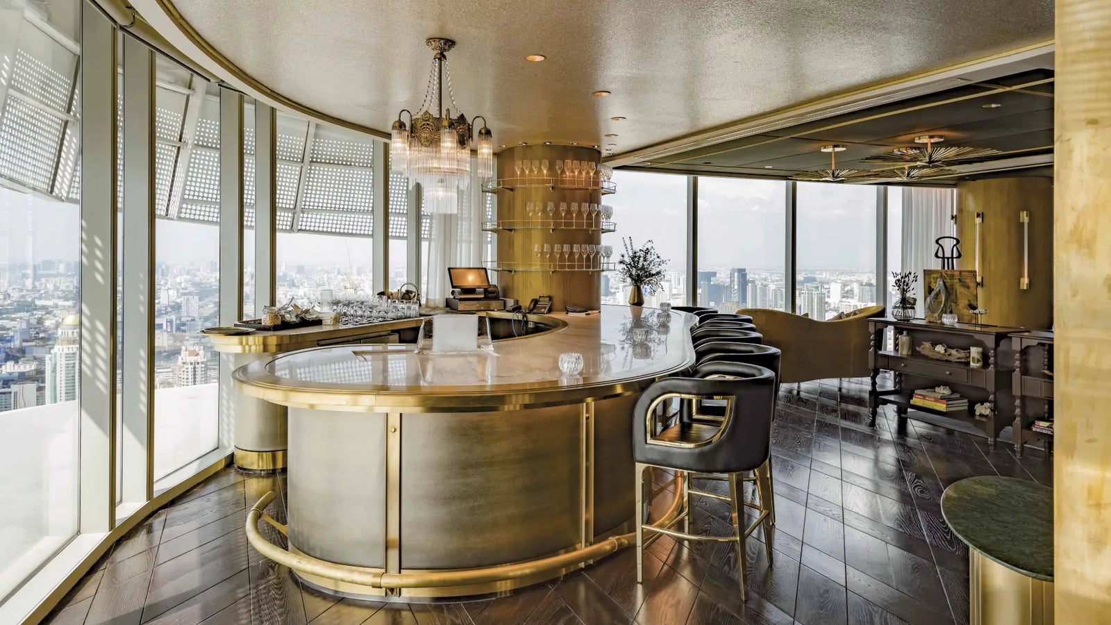 A luxury modern bar interior with a curved metallic bar counter, black bar stools, a glassware dispenser, and large windows overlooking a city skyline.