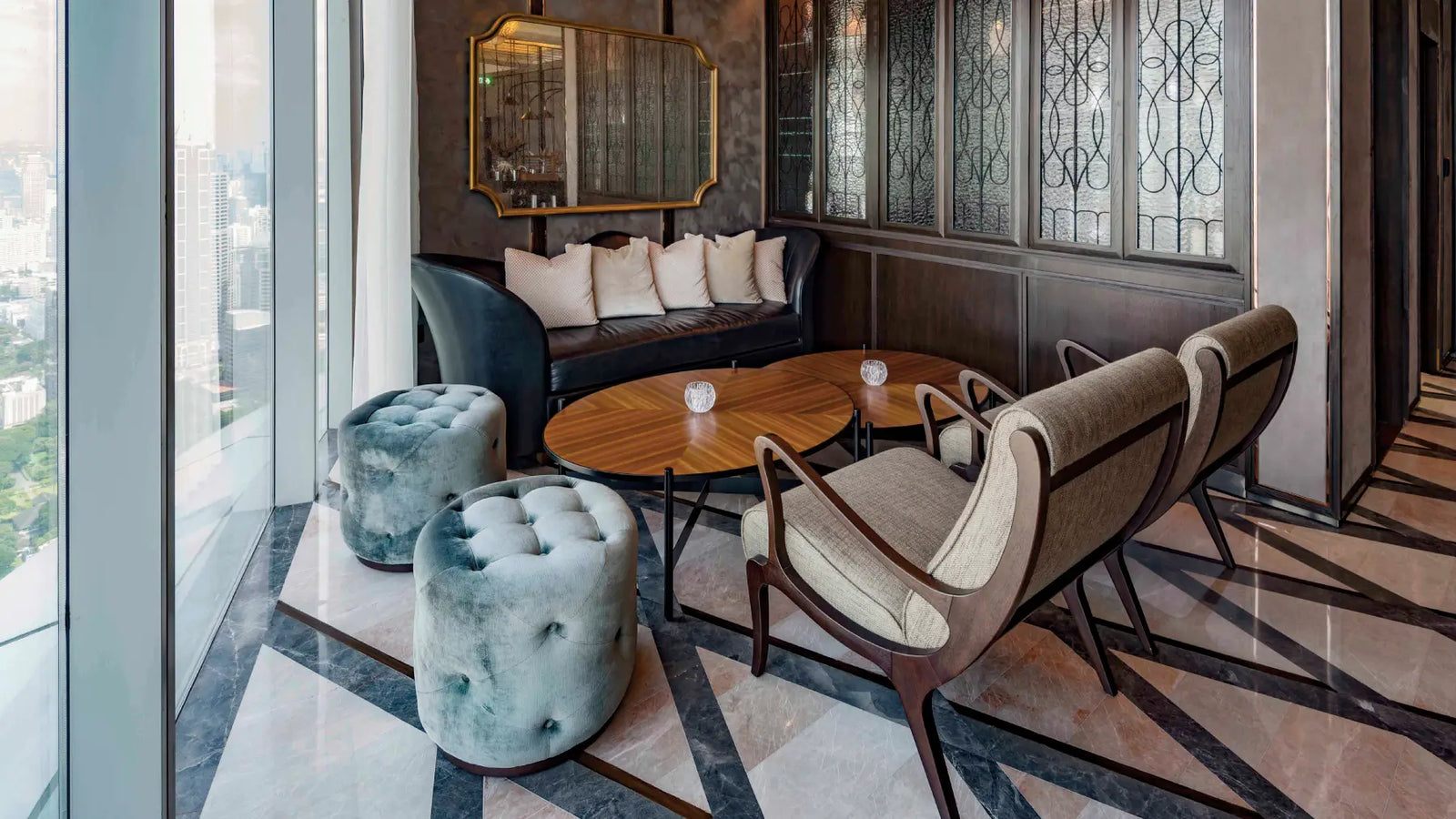 A luxury modern living room corner featuring two tufted velvet ottomans in a muted blue-green, two upholstered armchairs in beige and brown, and a dark leather sofa with cream pillows, arranged around a round wooden coffee table on a geometric marble floor.