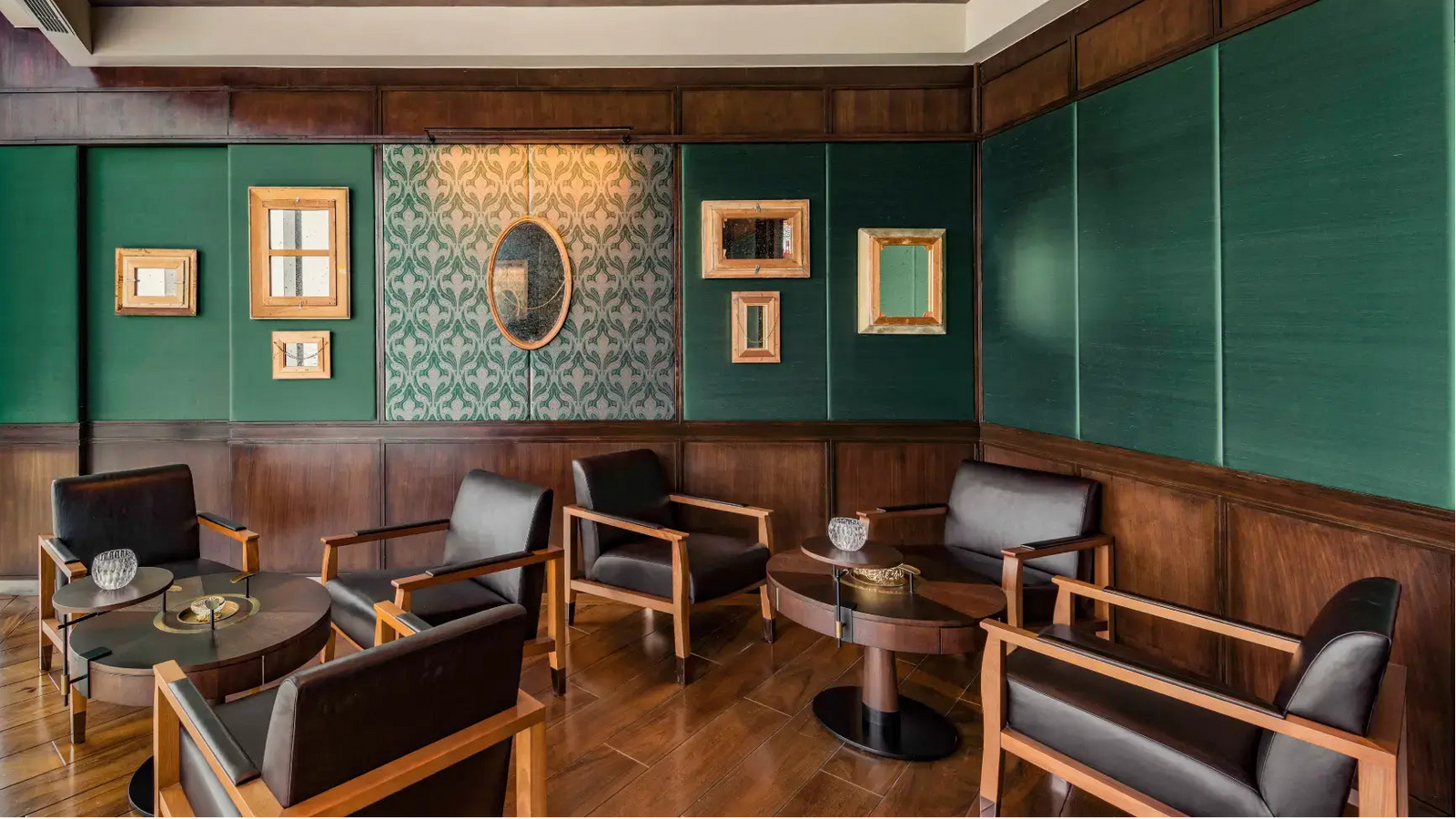 A luxury lounge or bar seating area with dark wood paneling, deep green accent walls, framed wall art, and multiple black leather armchairs arranged around small round wooden tables.