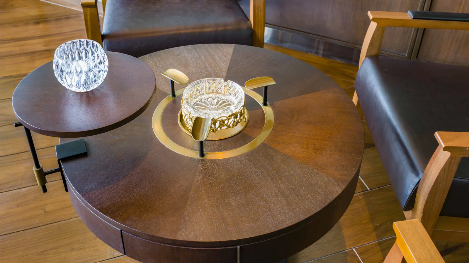 A round dark wood coffee table with a gold-toned central ring and a clear glass bowl centerpiece, shown in a furnished room with leather chairs and wood flooring.