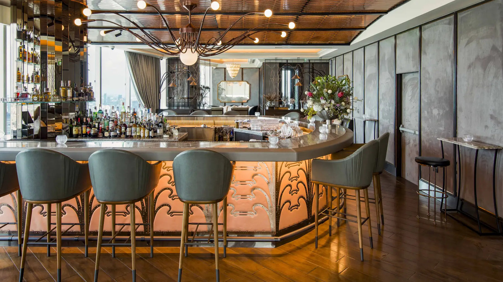 A modern bar interior with a curved counter, four gray bar stools, warm wood flooring, and a chandelier overhead. No people or animals are present.