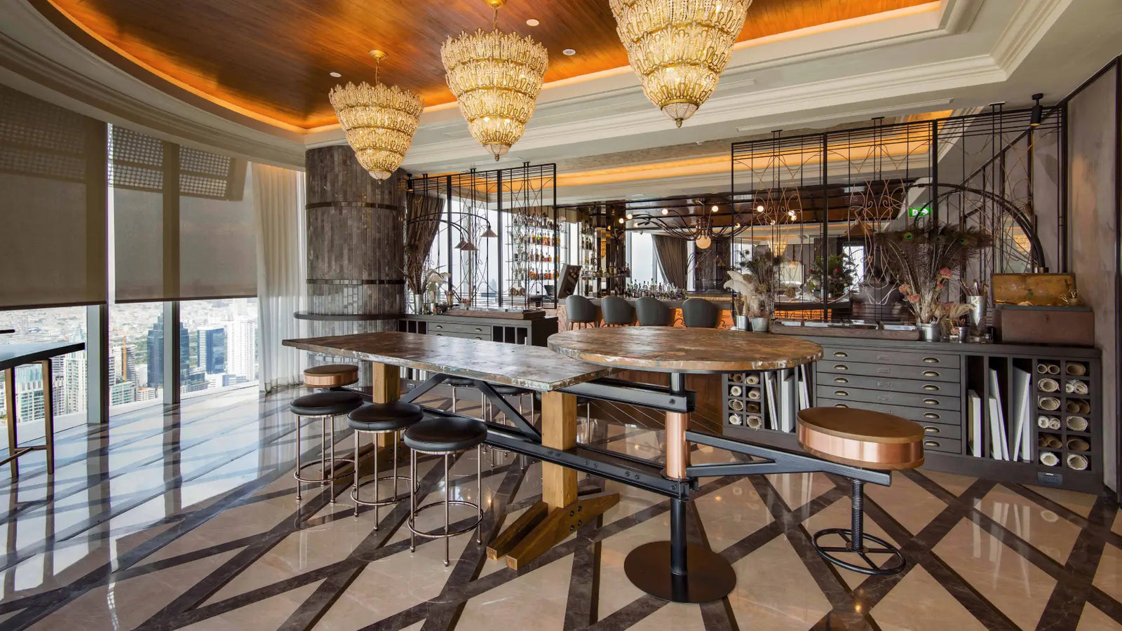A luxury modern bar and dining area with a long wooden bar table, black metal frames, and multiple bar stools, set in a high-end interior with chandeliers, a tiled floor, and a city view.