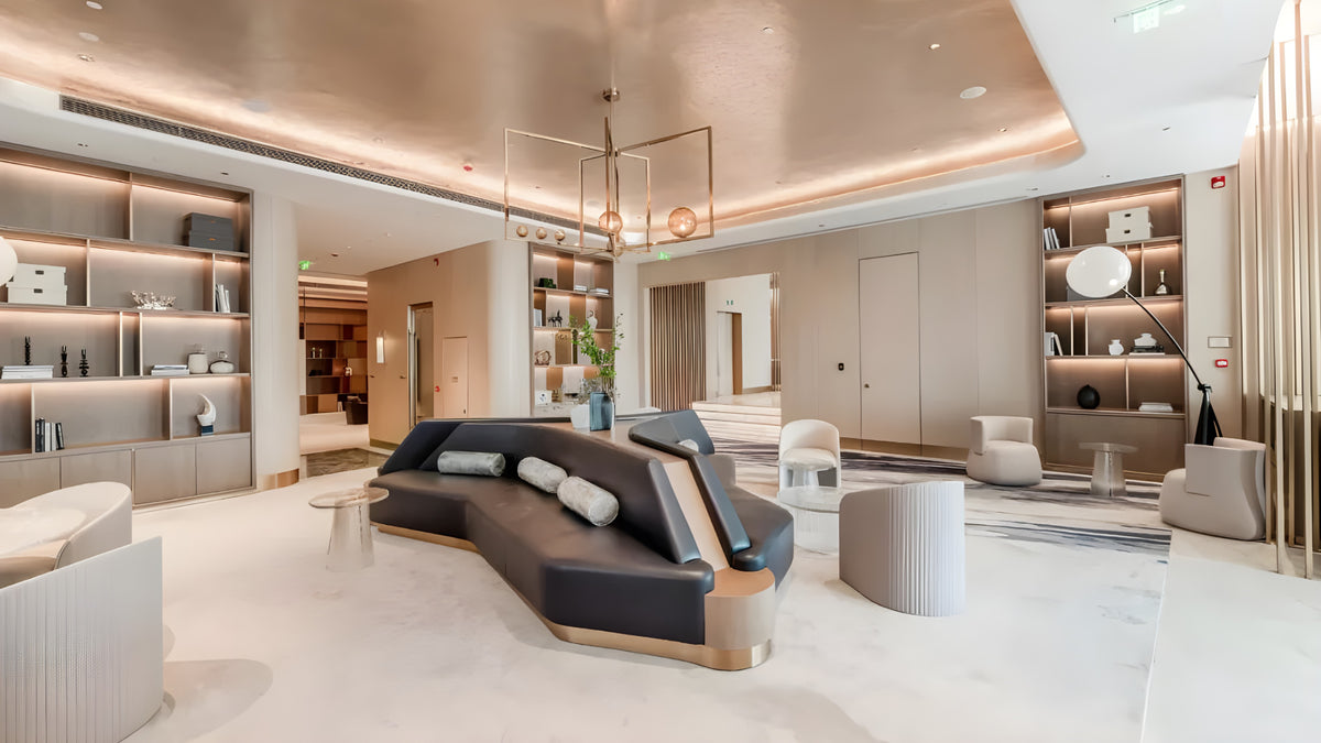 A modern lounge area featuring a curved dark gray sofa with light wood trim, ribbed white cylindrical ottomans, built-in shelving with warm lighting, and a spacious neutral-toned interior.