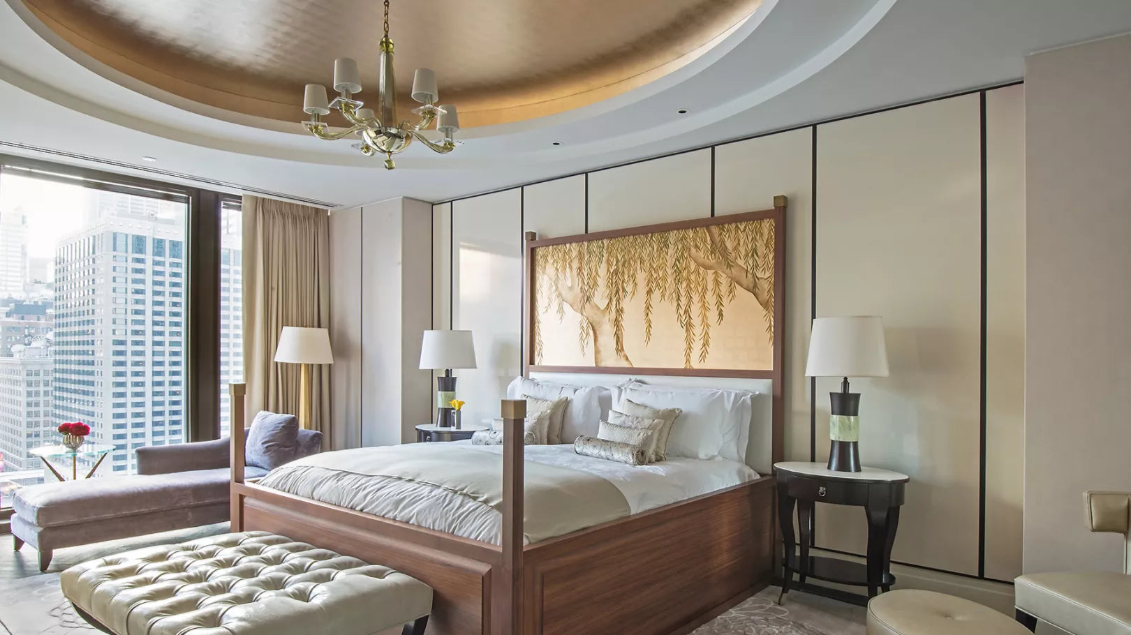 A luxury bedroom interior featuring a wooden four-post bed with white linens, a large framed wall art piece depicting hanging branches, a tufted bench, bedside tables with lamps, and a modern circular chandelier in a high-rise city apartment.